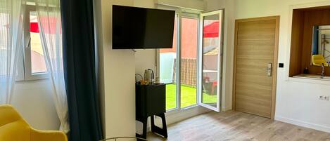 Una habitación moderna con televisión, una mesita, una silla amarilla y un ventanal corredero que da a un jardín.