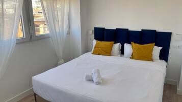 A neatly made bed with white linens and two yellow pillows, a blue headboard, and a window with sheer curtains.