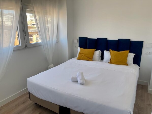 Una cama bien hecha con sábanas blancas y dos almohadas amarillas, un cabecero azul y una ventana con cortinas traslúcidas.