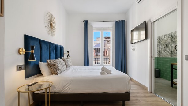 A modern bedroom with a large bed, a bedside table, a wall-mounted TV, and a view of the city through a window. - Hotel Málaga City Suites (Málaga)