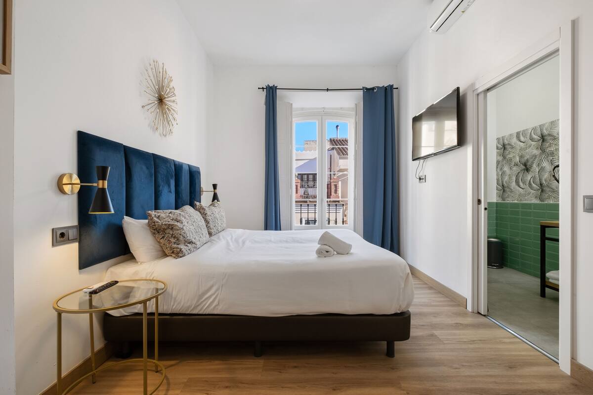 Ein modernes Schlafzimmer mit einem großen Bett, einem Nachttisch, einem an der Wand befestigten Fernseher und Blick auf die Stadt durch ein Fenster.