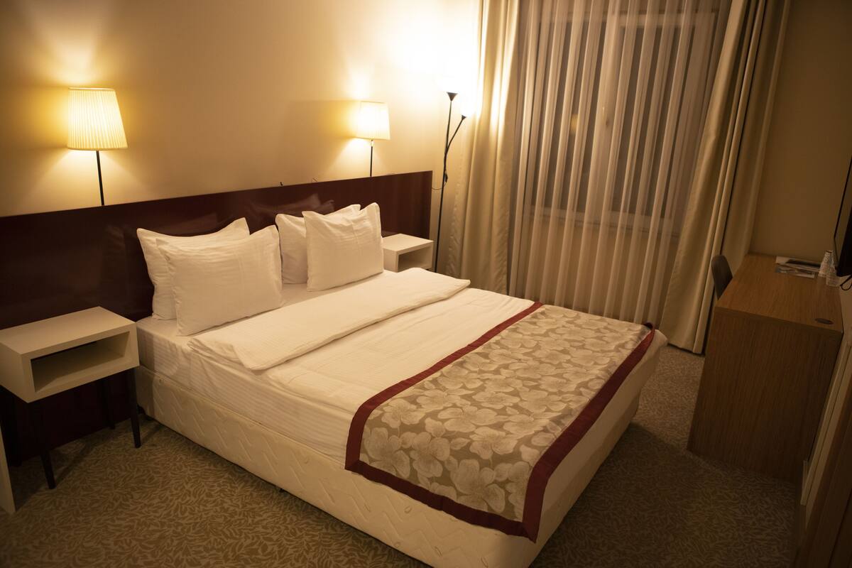 A hotel room with a bed, two bedside lamps, a nightstand, a wooden desk, and a window with curtains.