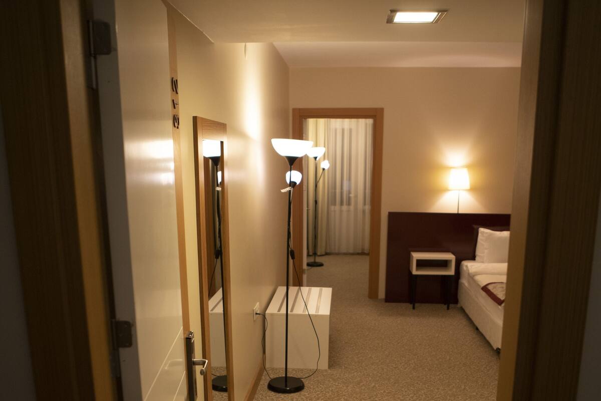 A hotel hallway with a carpeted floor, a bedside table with a lamp, a bed, and a mirror on the wall.