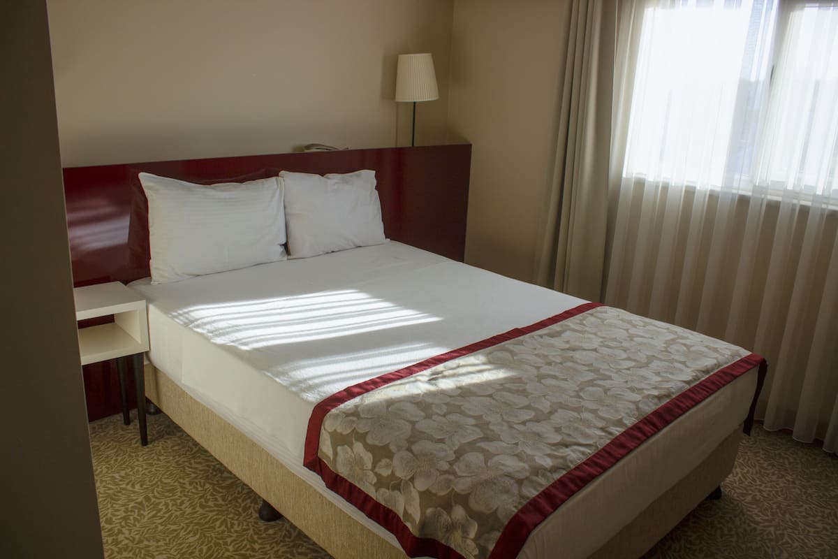 A neatly made bed with a floral-patterned bedspread and a red border, a bedside table with a lamp, and a window with sheer curtains allowing natural light.