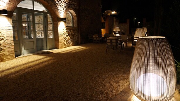 An outdoor area with a stone building, a door, and several chairs and tables under warm lighting. - Can Ribalta Hotel Gastronòmic (Sant Antoni de Vilamajor)