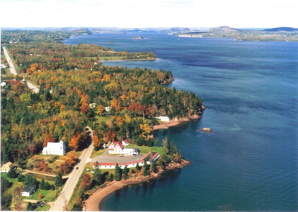 An aerial view of a coastal area with a prominent white building surrounded by autumn foliage, a road, and a large body of water.