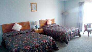 Quarto de hotel com duas camas, mesa de cabeceira com abajur, uma cadeira e janela com cortinas.