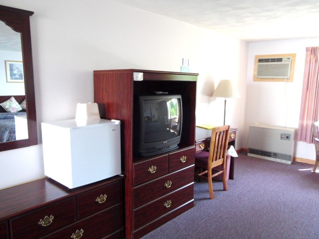 Une chambre d’hôtel avec un meuble en bois pour la télévision, un mini-réfrigérateur blanc, un bureau avec une lampe et une chaise.