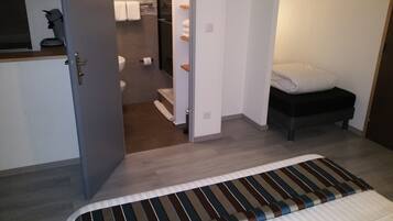 Una habitación de hotel con cama, un pequeño banco, un inodoro y una zona de ducha.