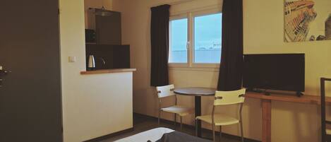 A hotel room with a bed, a desk with a television, a chair, a small table, and a window with curtains.