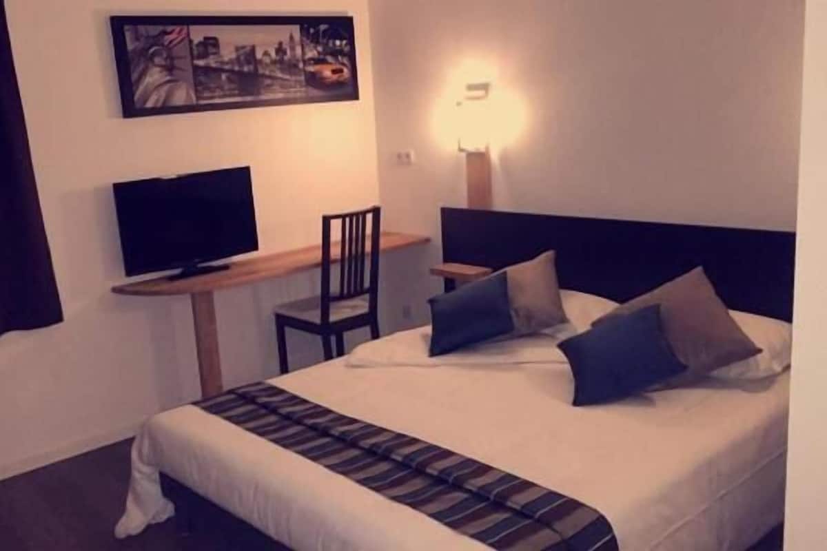 A hotel room with a bed, a desk with a television, a chair, and a lamp.