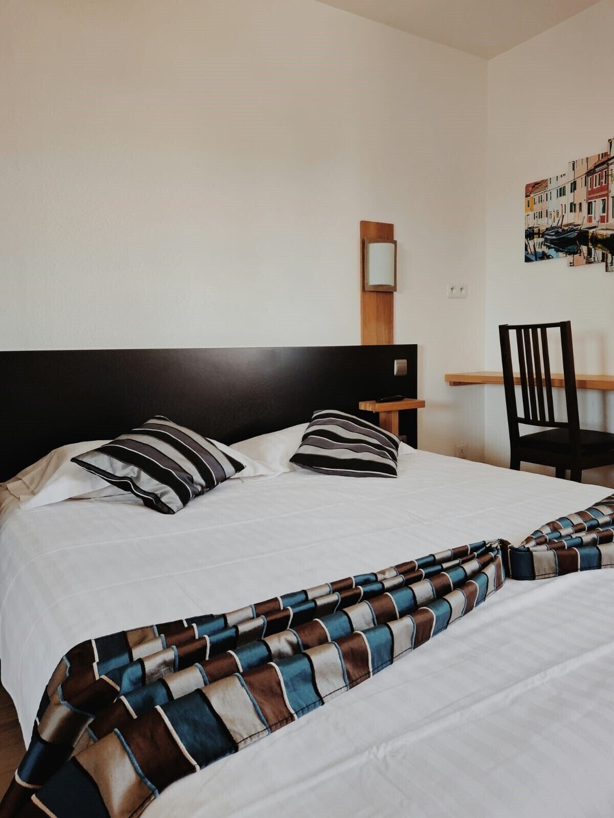 A neatly made bed with striped pillows and a striped blanket, a wooden nightstand with a lamp, and a chair.