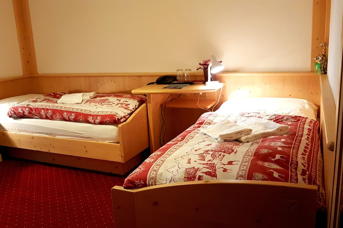 Une pièce avec deux lits en bois, chacun recouvert d’une couvre-lit à motifs rouges et blancs. On y trouve une petite table de chevet en bois, sur laquelle sont posés une lampe et quelques livres. On aperçoit également un bureau en bois avec une lampe et une petite plante.