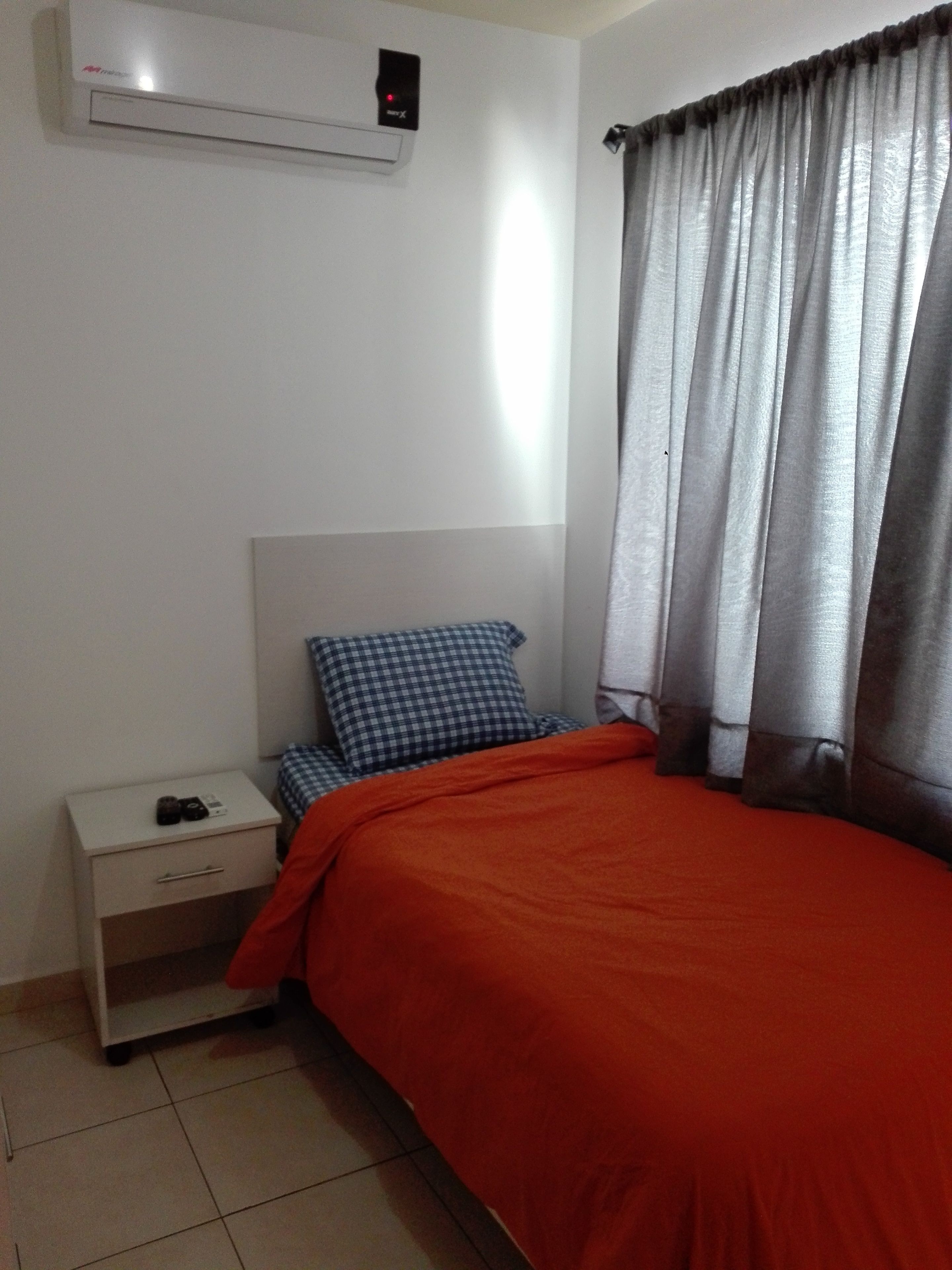 A bedroom with a bed, a nightstand, a checkered pillow, a red blanket, a grey curtain, and an air conditioning unit.