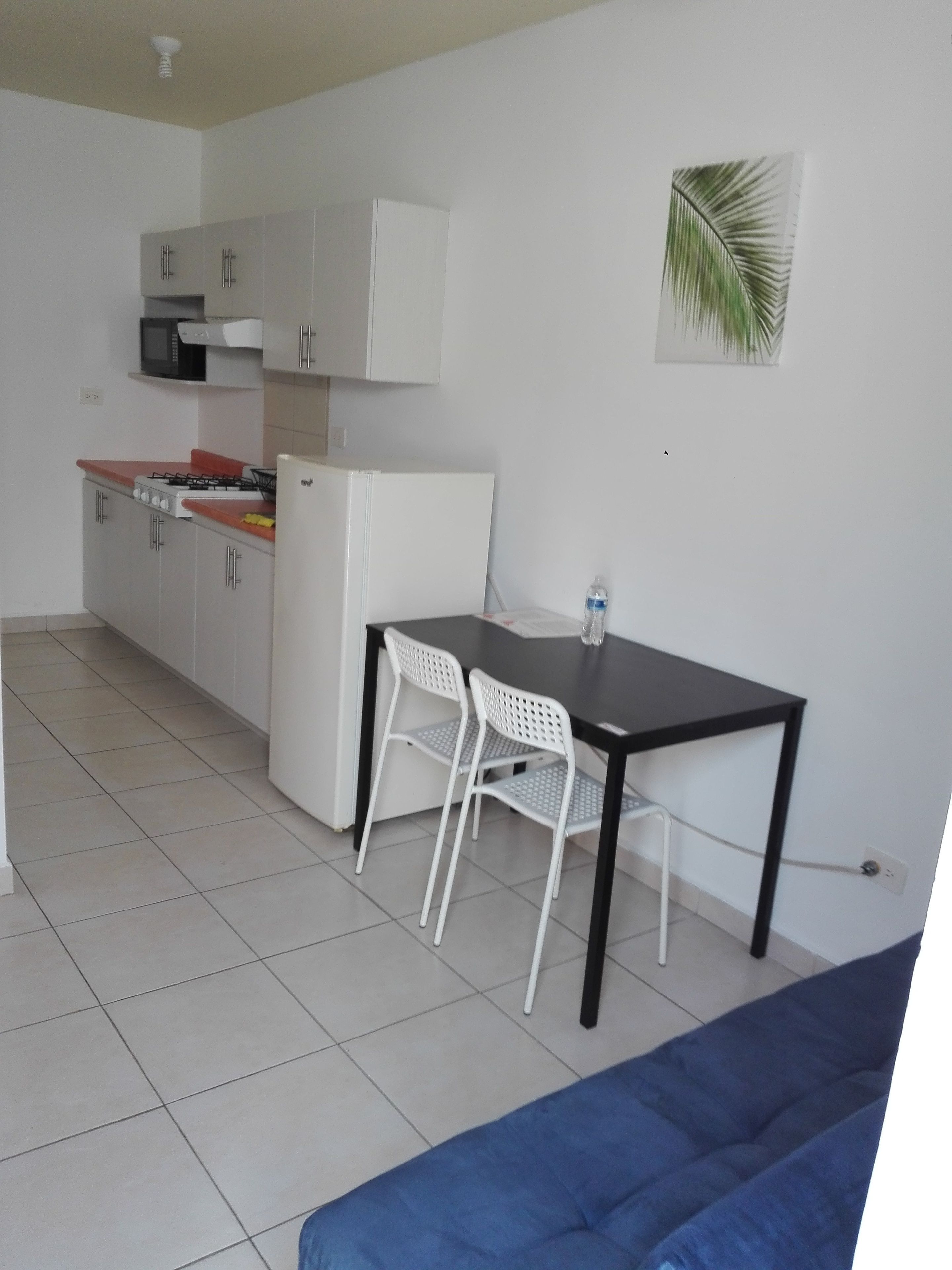 Una cocina compacta con armarios blancos, un fogón y un microondas. Se ve una mesa de comedor con dos sillas y un sofá azul al frente.