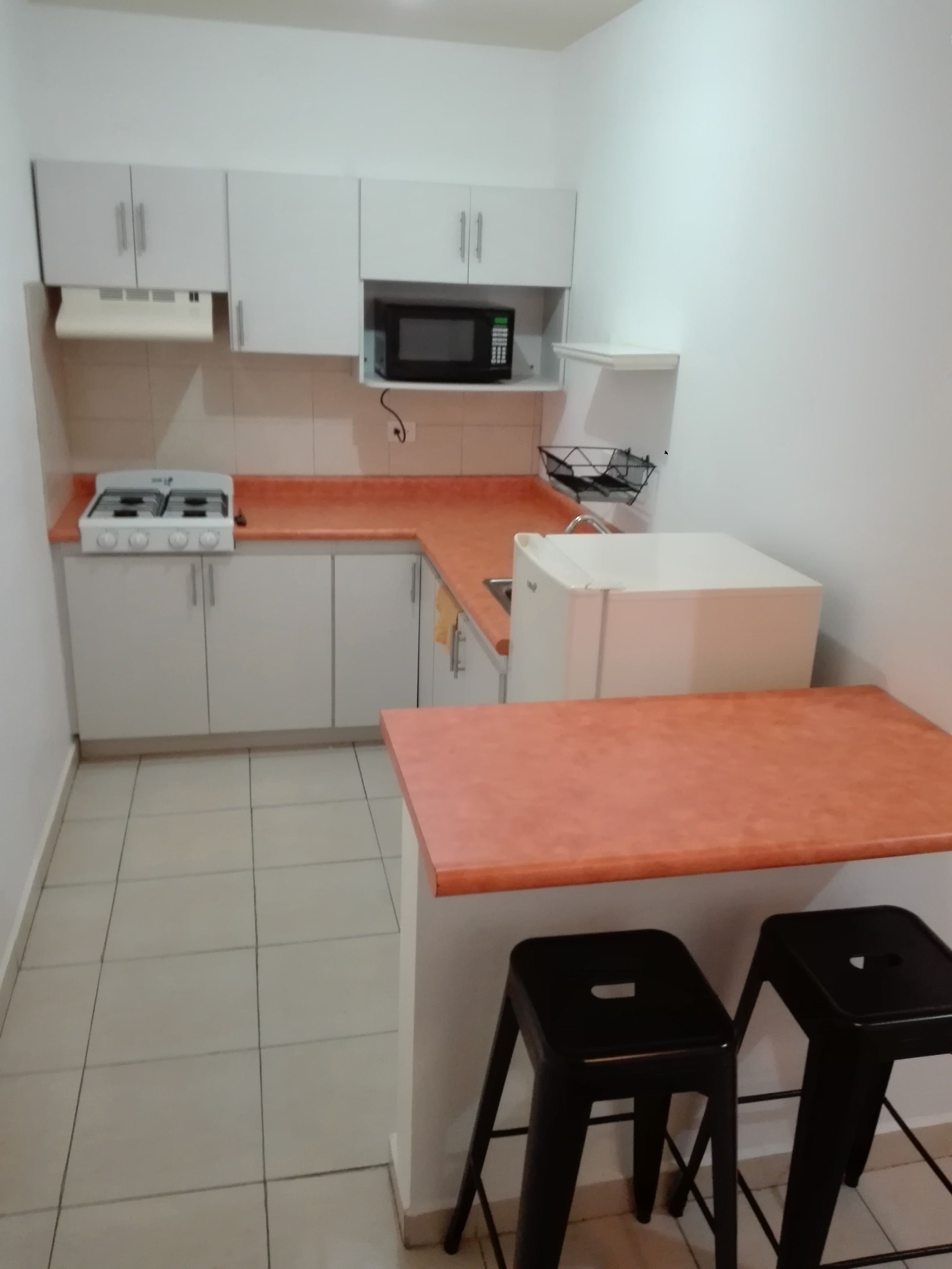 Una cocina funcional con armarios blancos, microondas, cocina a gas y dos taburetes negros.