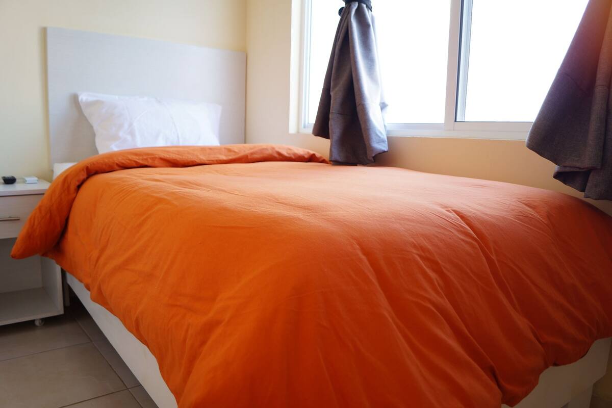 Una cama bien hecha con una colcha naranja, un cabecero blanco y una ventana con cortinas.