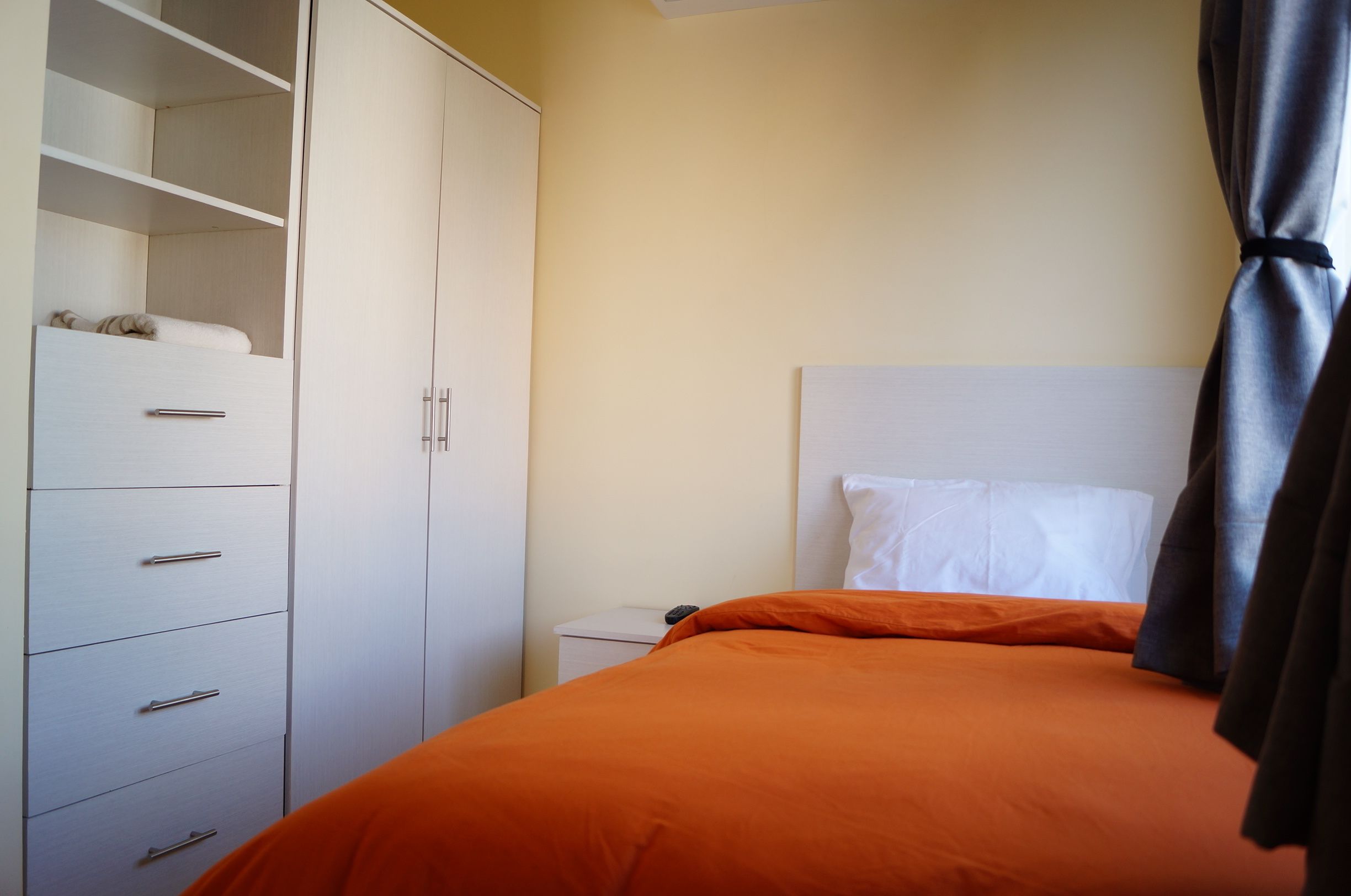 Un dormitorio con cama, colcha naranja, cabecero blanco, armario con varios cajones y mesita de noche con control remoto.