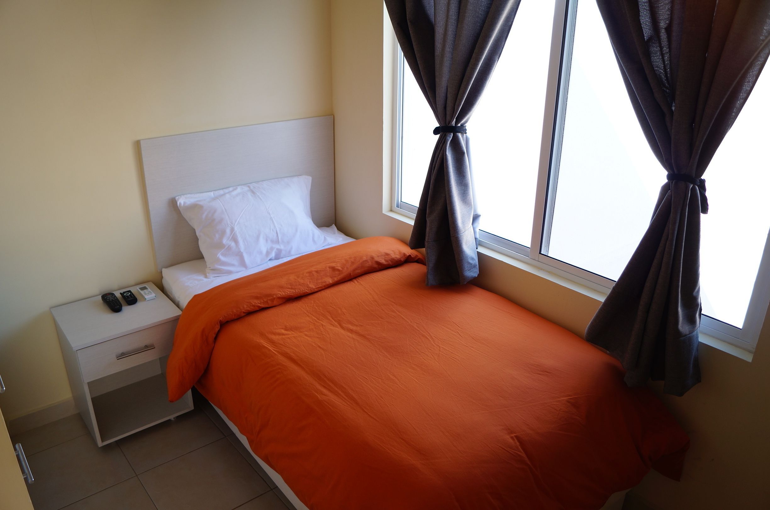 Una cama bien hecha con una colcha naranja, un almohadón blanco y cabecera blanca. Hay una mesita de noche con un control remoto y un cajón pequeño. La habitación tiene una ventana con cortinas oscuras recogidas.