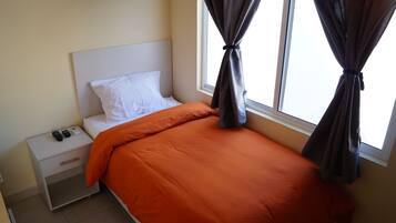 Una cama bien hecha con una colcha naranja, un almohadón blanco y cabecera blanca. Hay una mesita de noche con un control remoto y un cajón pequeño. La habitación tiene una ventana con cortinas oscuras recogidas.