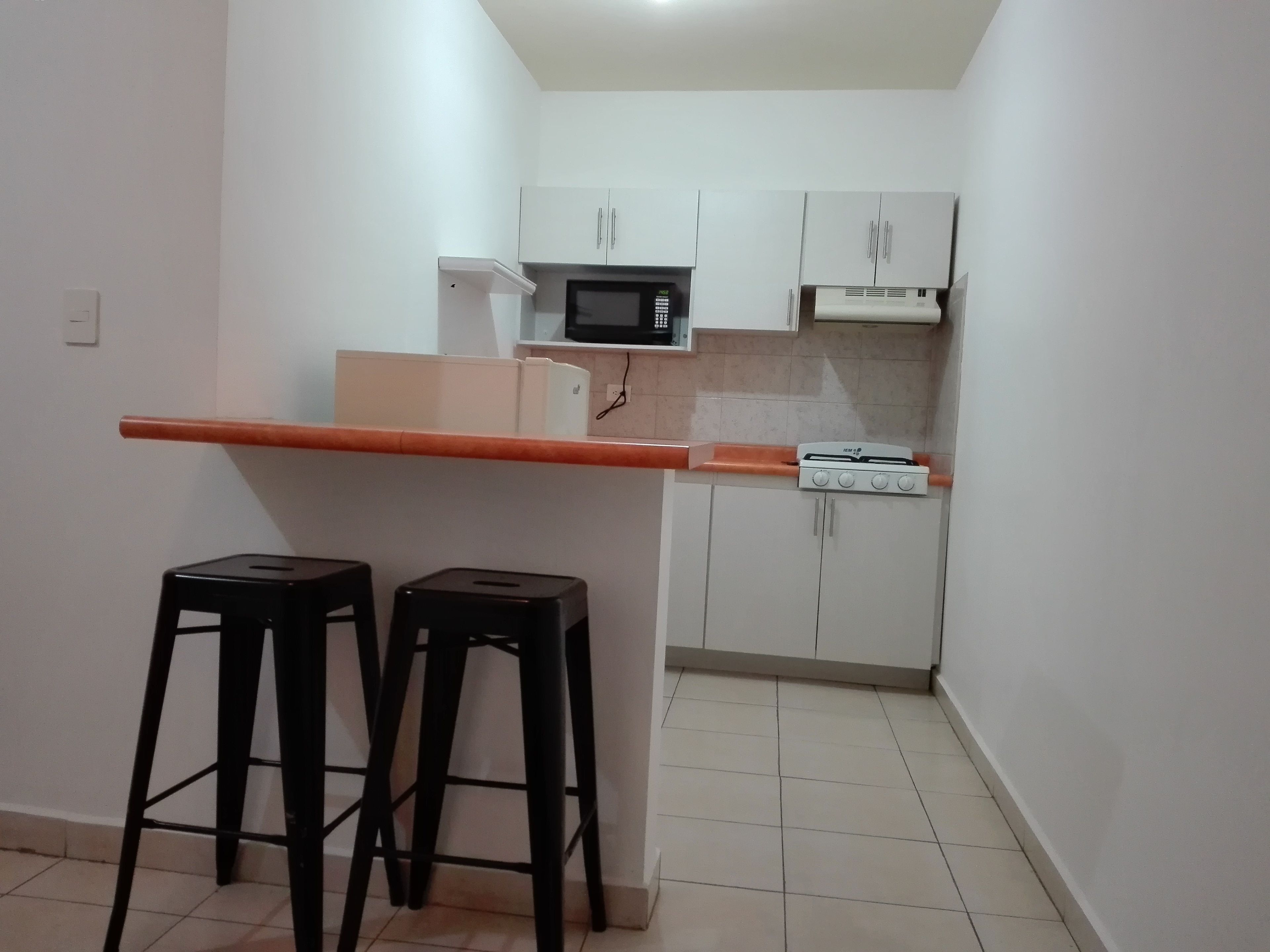 A small kitchen with white cabinets, a microwave, and a gas stove. There are two black stools and a countertop.