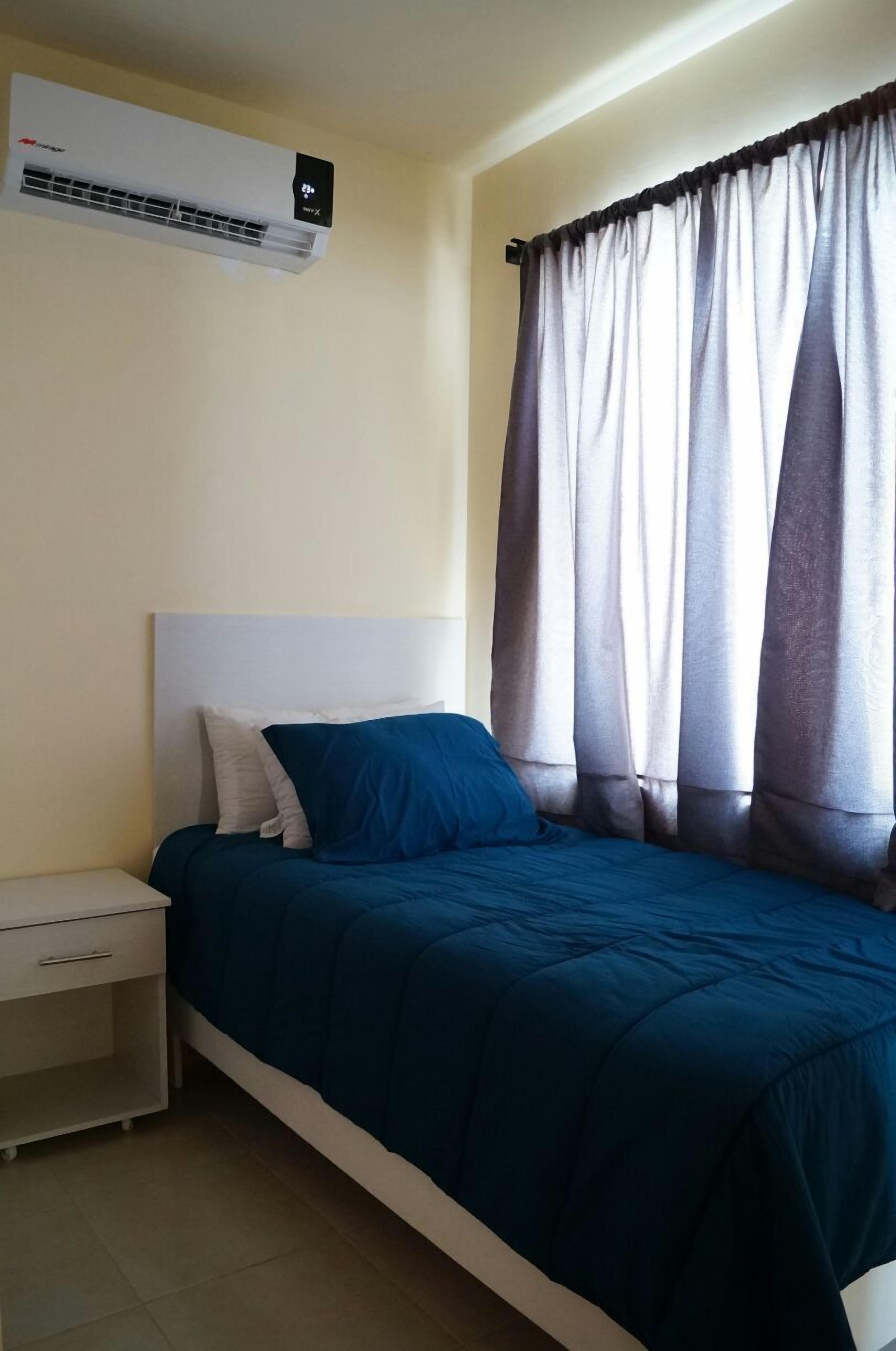 A bedroom with a bed, a nightstand, an air conditioner, and sheer curtains.