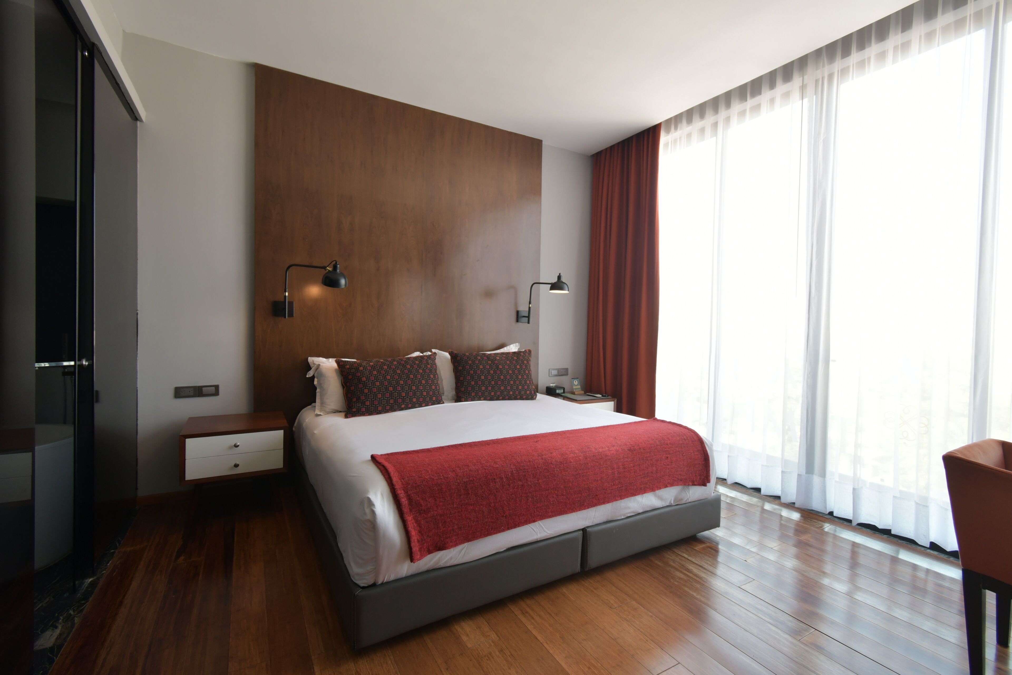 Un dormitorio moderno con una cama grande, un panel de pared de madera, dos lámparas de noche, una mesita de noche y un amplio ventanal con cortinas.