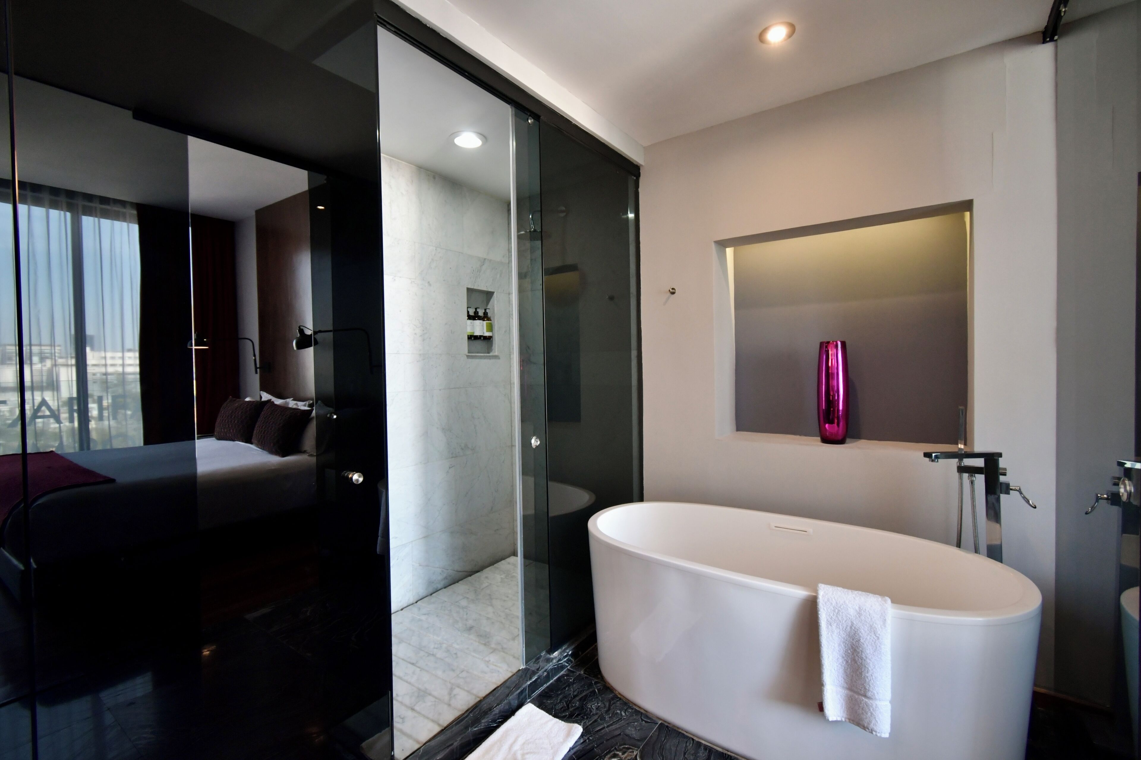 A modern bathroom with a large bathtub, a glass shower enclosure, and a bed with a view of the city.