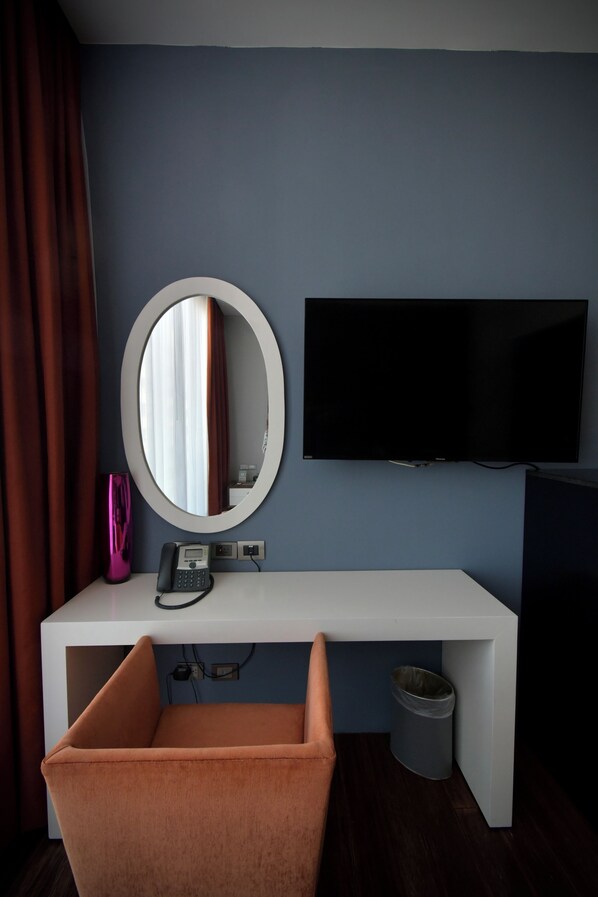 A modern hotel room with a white console table, a round mirror, a flat-screen TV, a telephone, and a trash bin.
