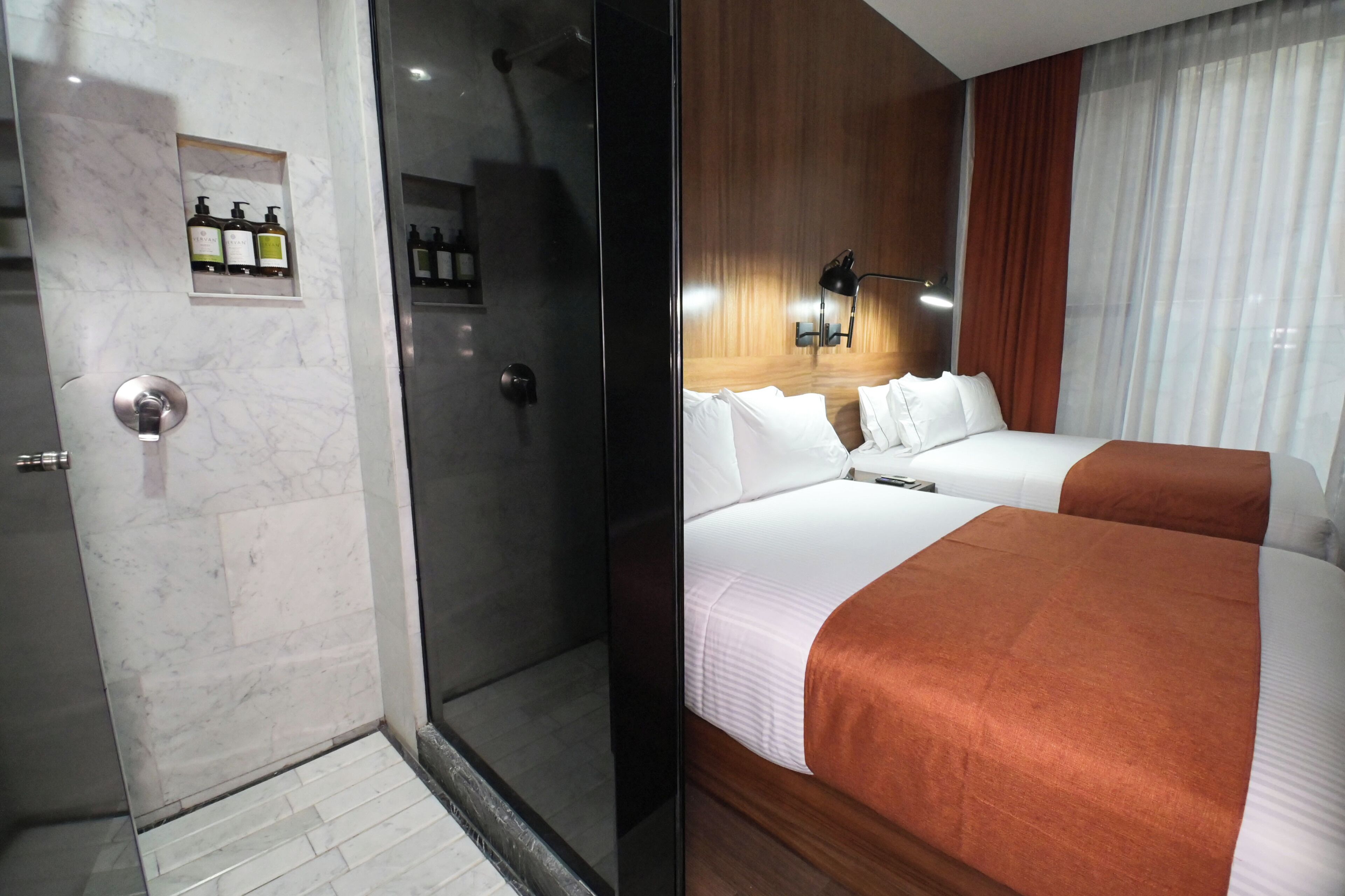 Ein Hotelzimmer mit Duschkabine aus Glas, einer Minibar mit Pflegeprodukten, einem Bett mit weißer Bettwäsche und einer braunen Tagesdecke sowie einem Holzkopfschild.