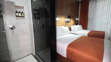 Habitación de hotel con cabina de ducha de vidrio, minibar con artículos de aseo, cama con ropa blanca y un plaid marrón, y cabecera de madera.