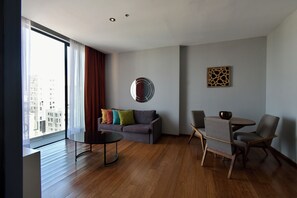 A modern living room with a grey sofa, colorful cushions, a round mirror, and a dining area with two chairs and a table. - Vivé Hotel Love it - CAS (Guadalajara)