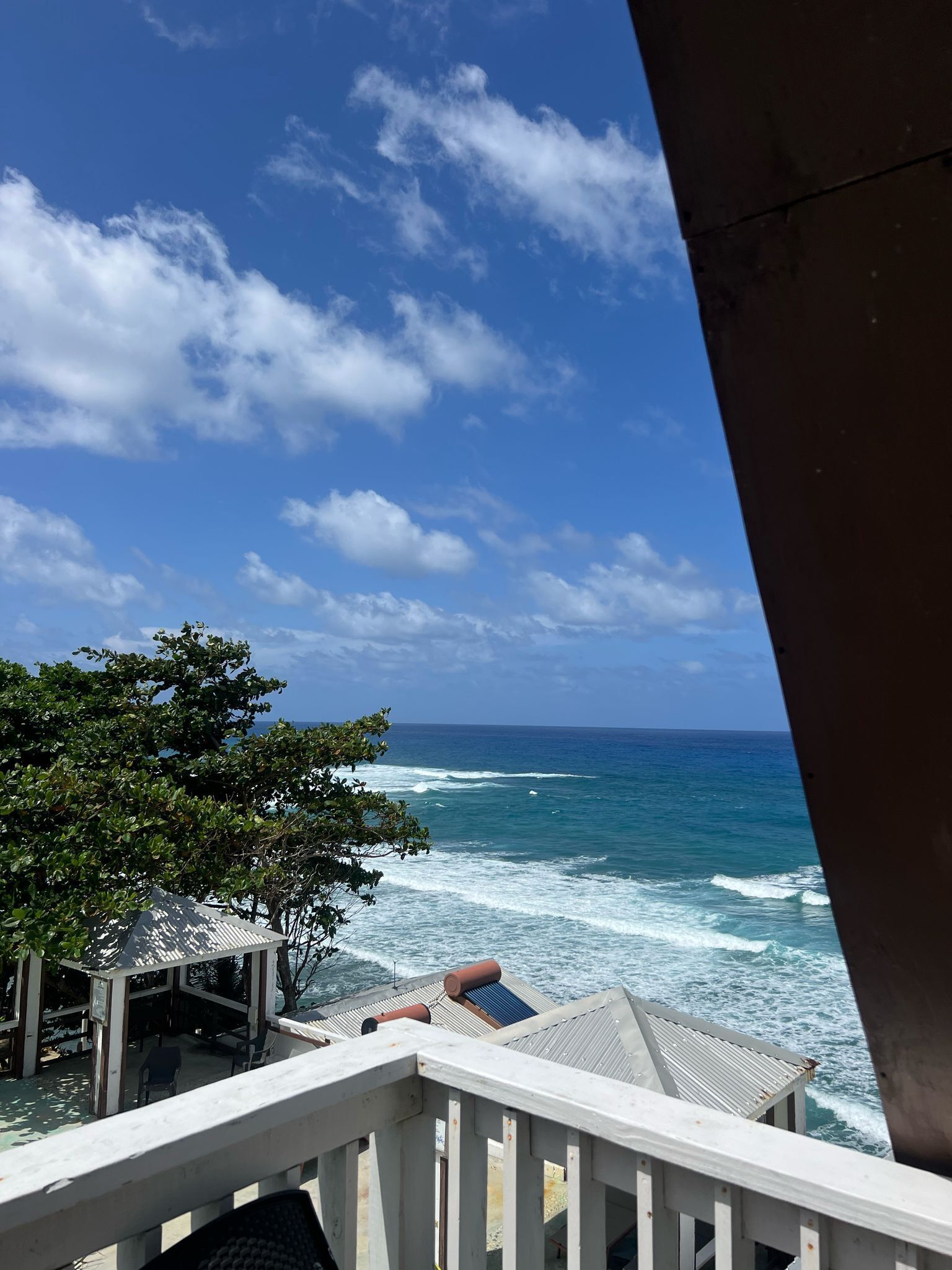 A view from a balcony overlooking the ocean with waves crashing onto the shore, a tree, and a building with a roof and railing.
