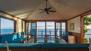 Una habitación amplia con un gran ventanal con vistas al océano, ventilador de techo y zona de estar con cojines azules.