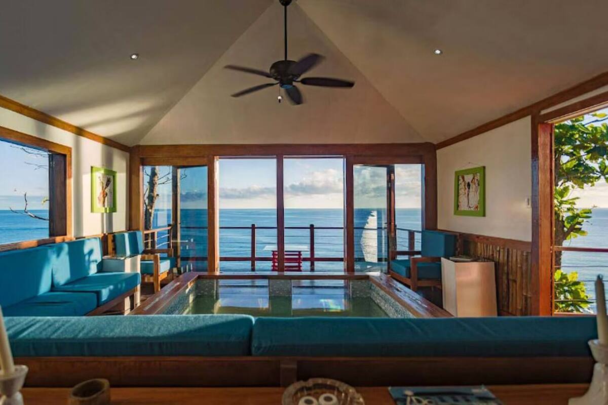 Una habitación amplia con un gran ventanal con vistas al océano, ventilador de techo y zona de estar con cojines azules.