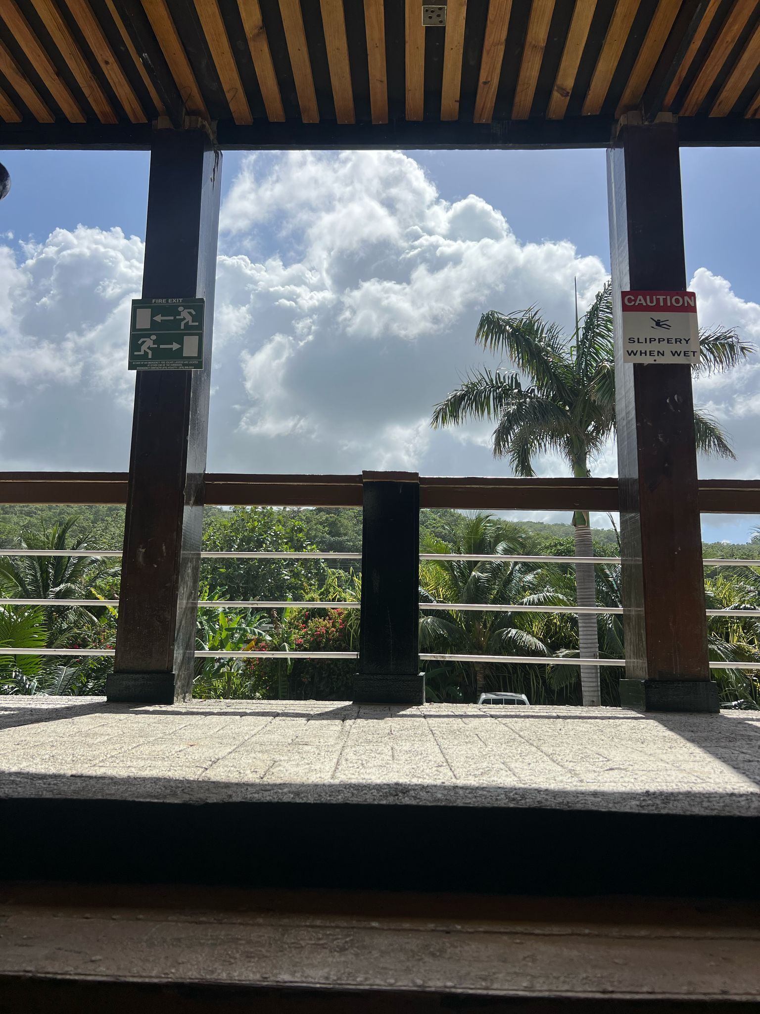A covered outdoor area with a fire exit sign, a caution sign about slippery surfaces when wet, and a view of palm trees and greenery.