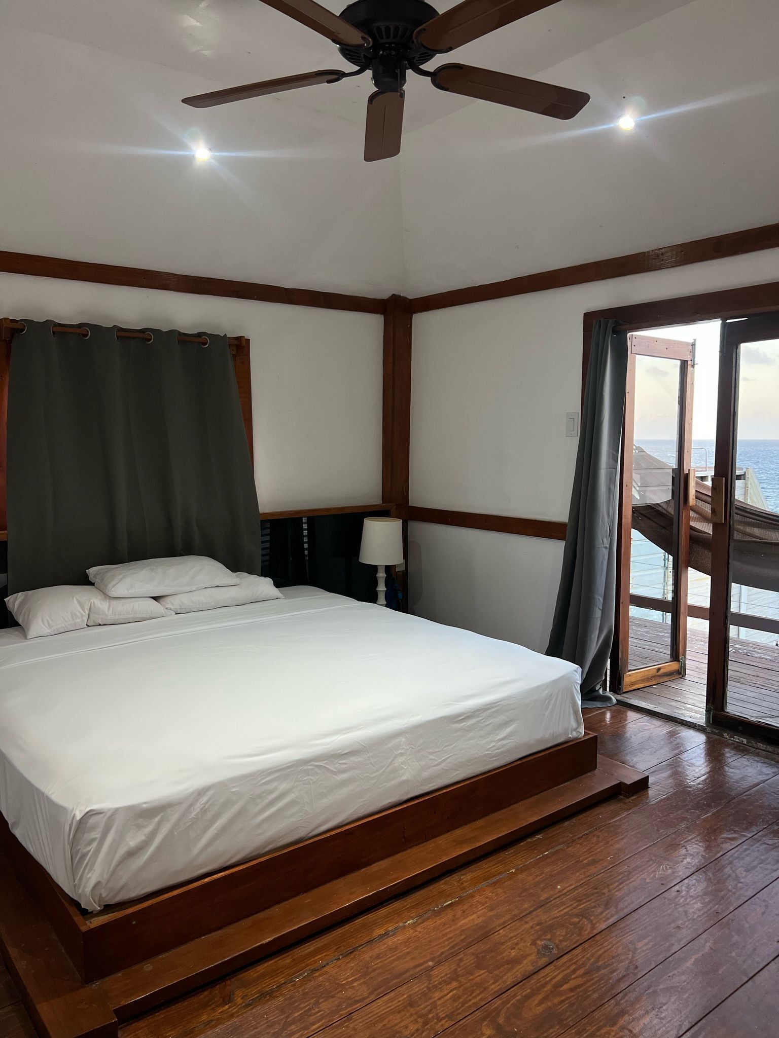A bedroom with a bed, a ceiling fan, a window with curtains, and a view of the sea.