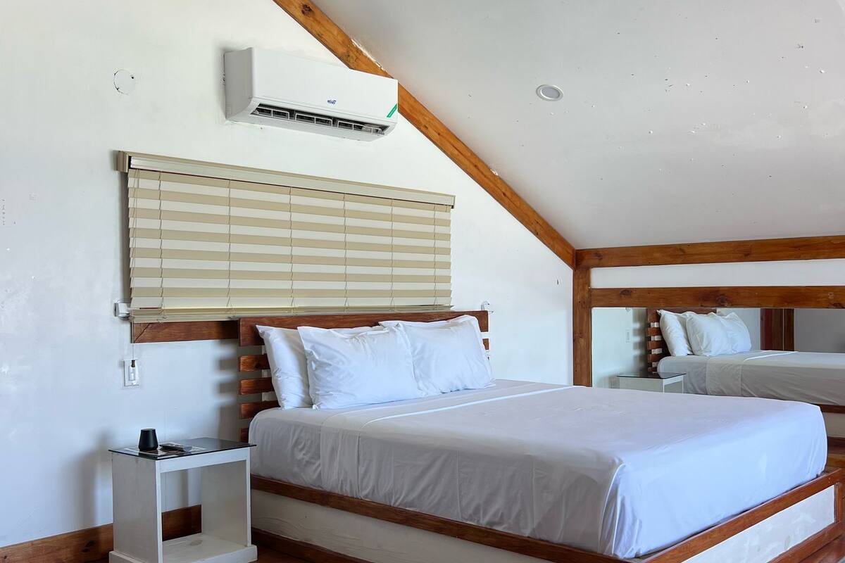 Un dormitorio con techo inclinado, ventilador de techo, cama con ropa de cama blanca, mesita de noche y ventana con persianas.