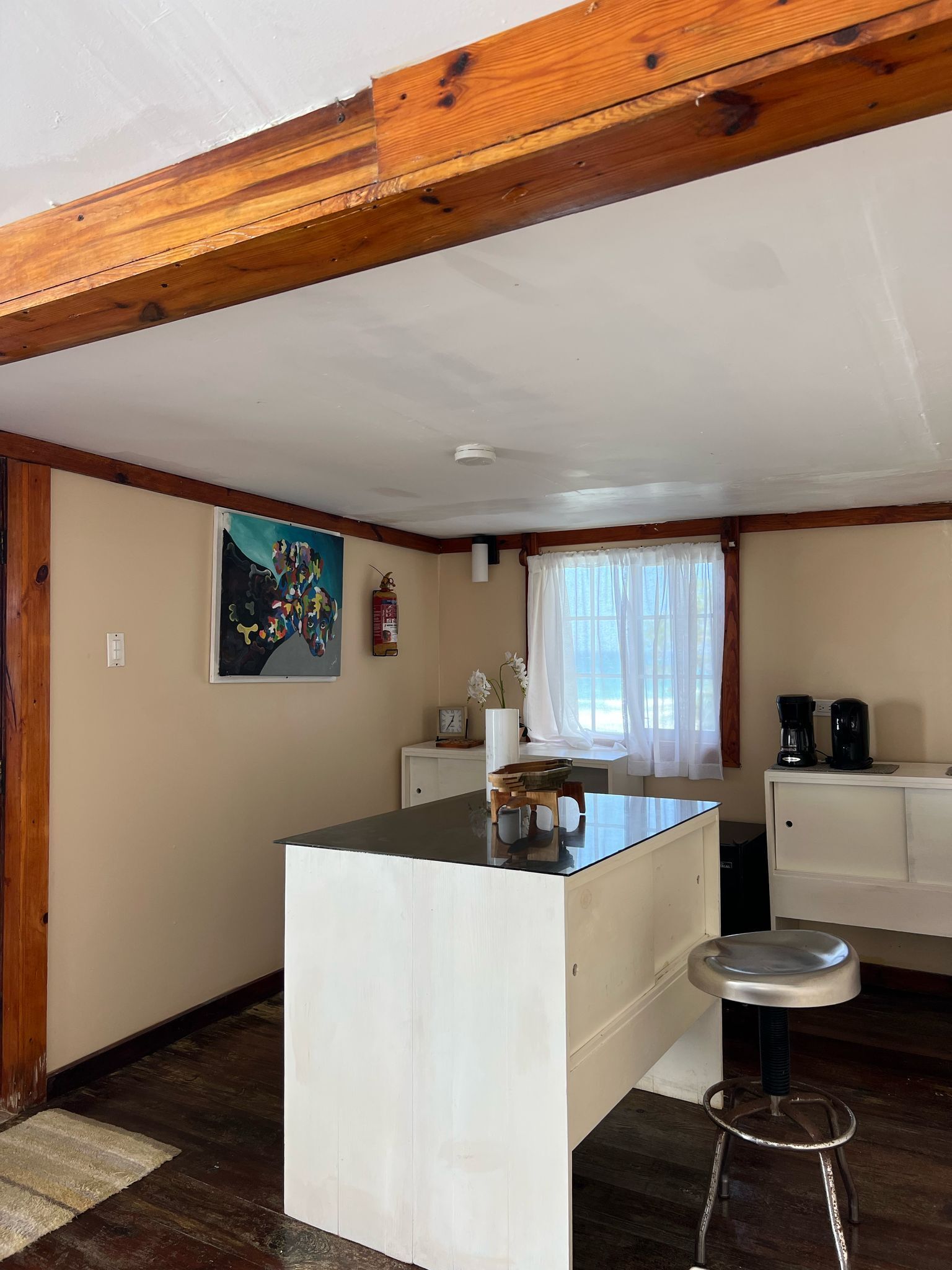A kitchen with a central island, wooden beams, a white cabinet, a coffee maker, and a window with curtains.
