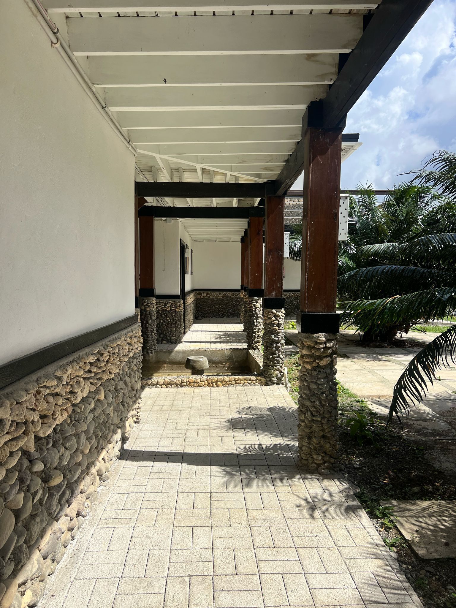 A covered walkway with stone pillars and a tiled floor, leading to a building entrance.