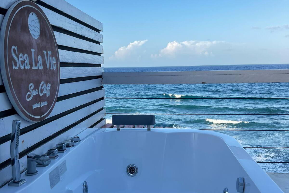 Tina de hidromasaje blanca con un letrero que dice "Sea La Vie Sea Cliff" frente a una playa y el océano.