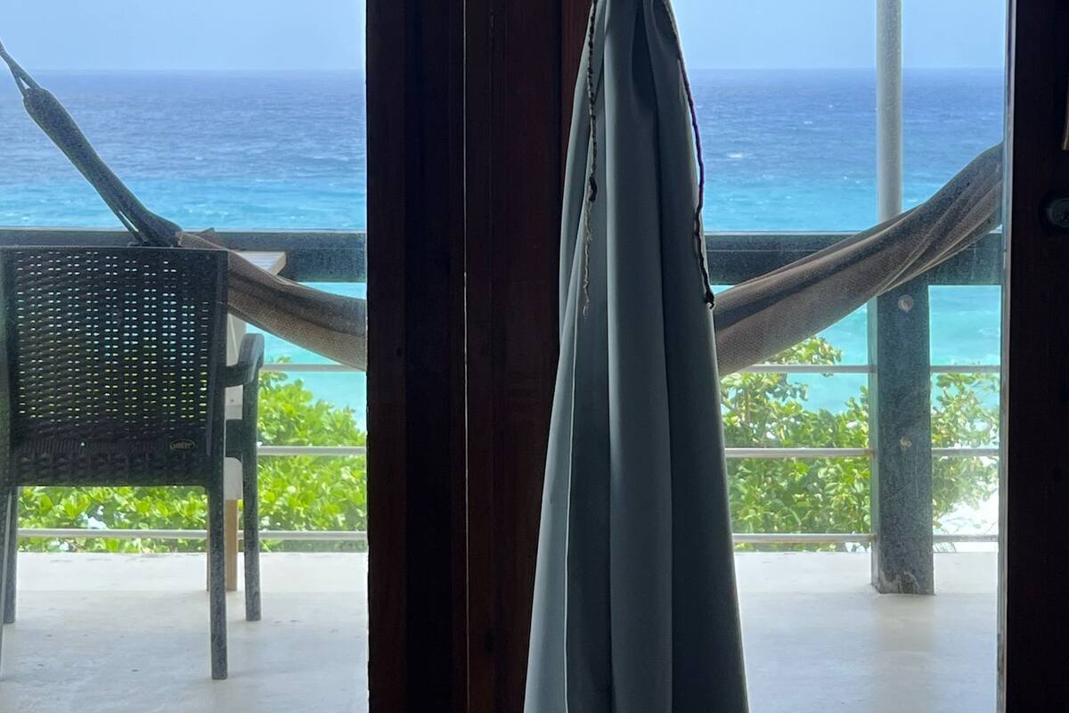 Vista desde una habitación con un ventanal grande, que incluye una hamaca y una silla en el exterior, con vista al océano.