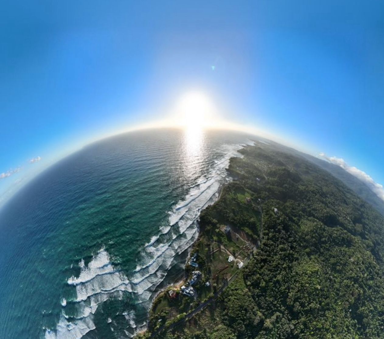 An aerial view of a coastal area with a clear sky, ocean waves, and a forested landscape.
