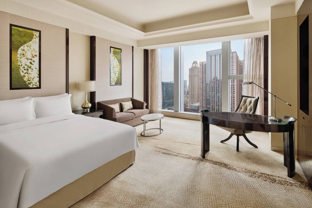 A hotel room with a large bed, a sofa, a small round table, a desk, and a view of tall buildings through large windows.