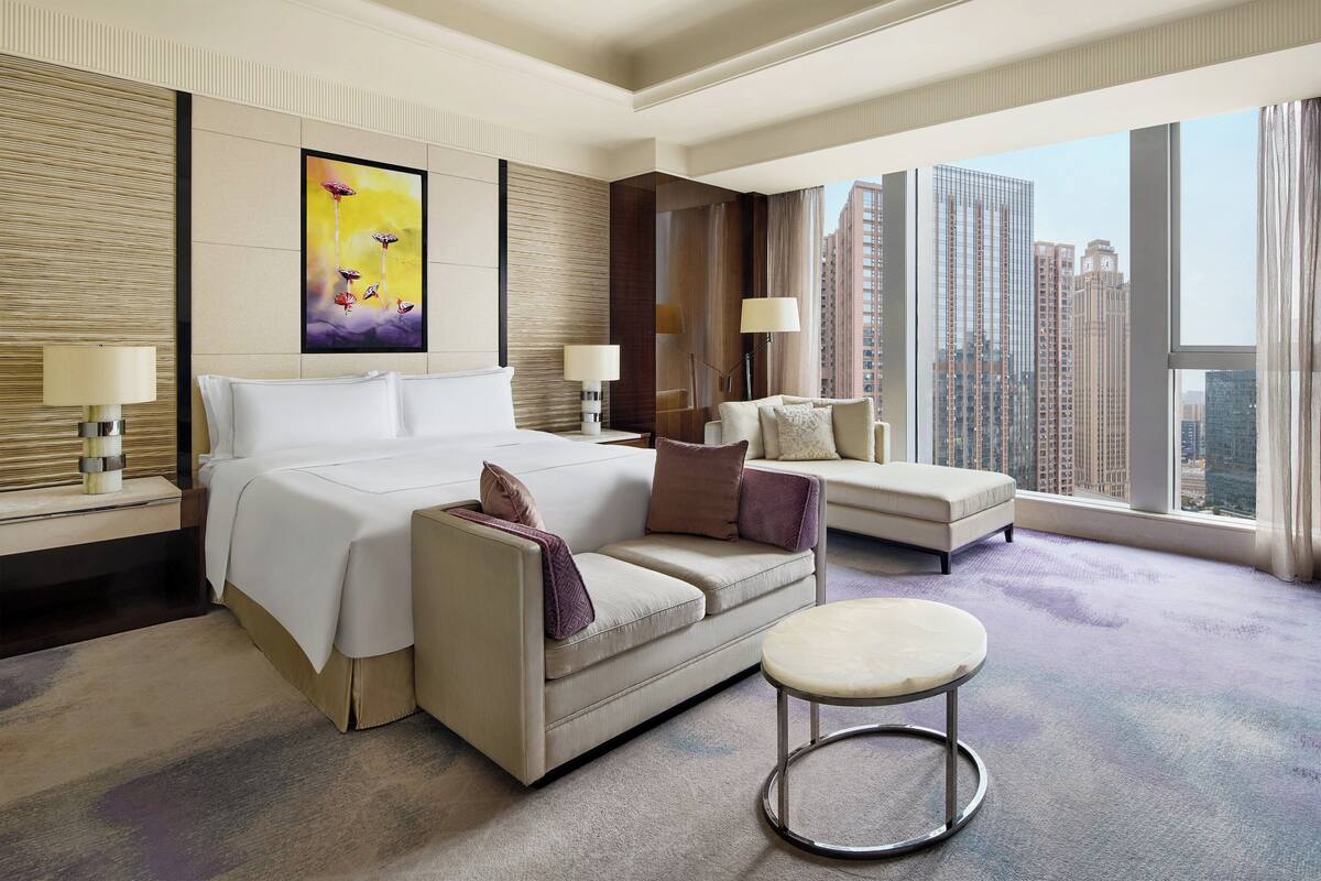 A modern hotel room with a large bed, a sofa, a small table, and a city view.