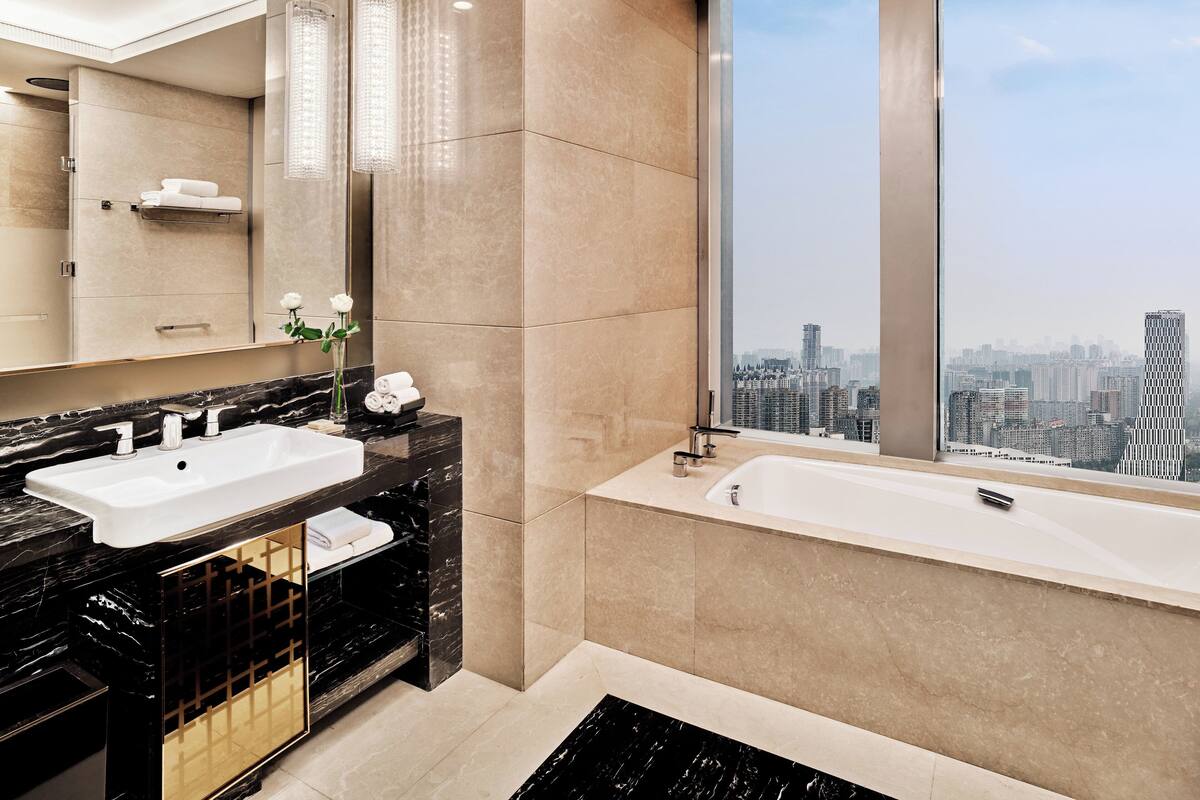 A modern bathroom with a large mirror, two sinks, a bathtub, and a city view through a large window.