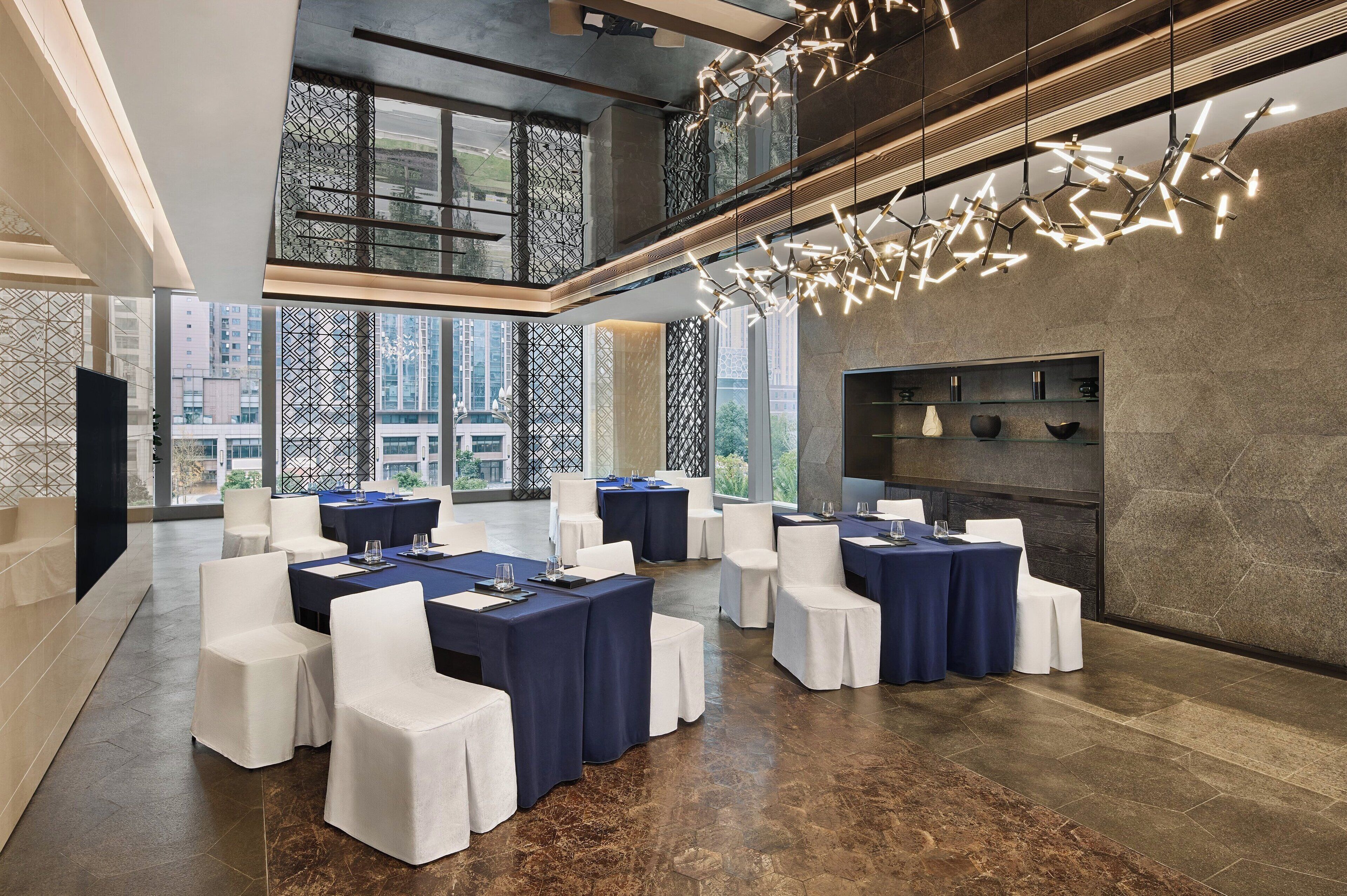 A spacious conference room with high ceilings, large windows, and a modern chandelier. Tables are set with white chairs and blue tablecloths.