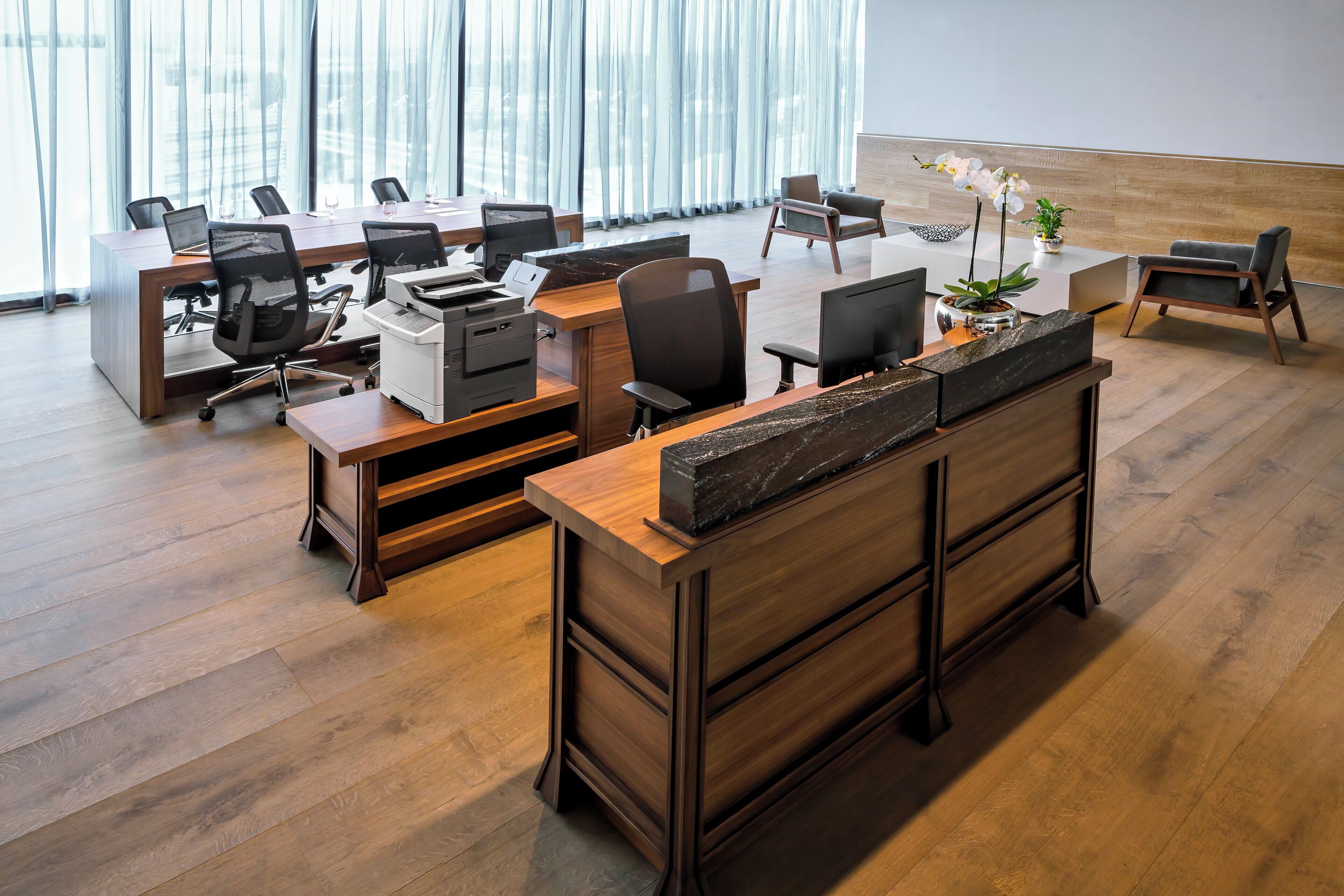 A modern office space with wooden desks, black office chairs, and a reception desk with a plant.