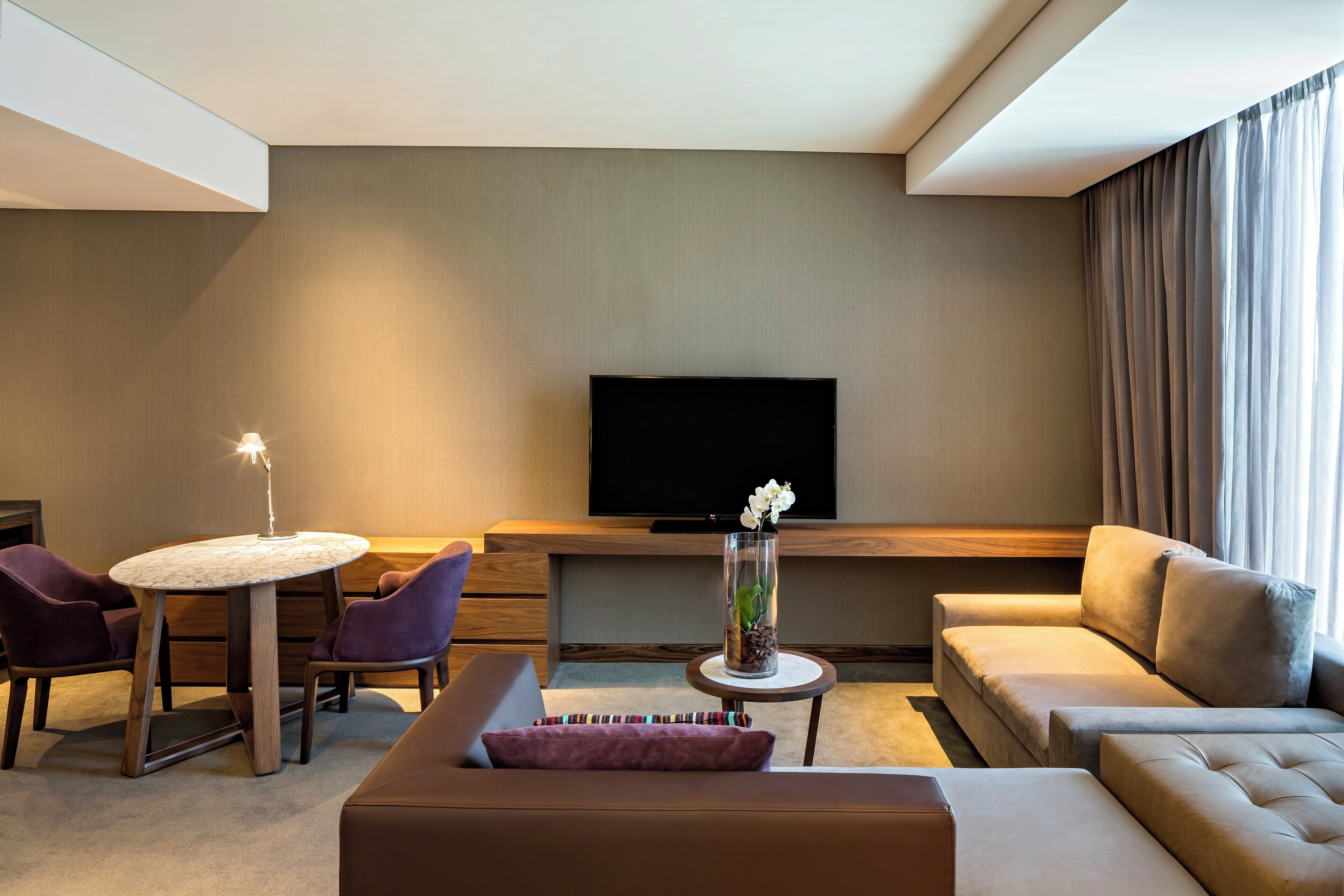 A modern hotel room with a flat-screen TV, a round dining table with chairs, a sofa, and a decorative vase with flowers.