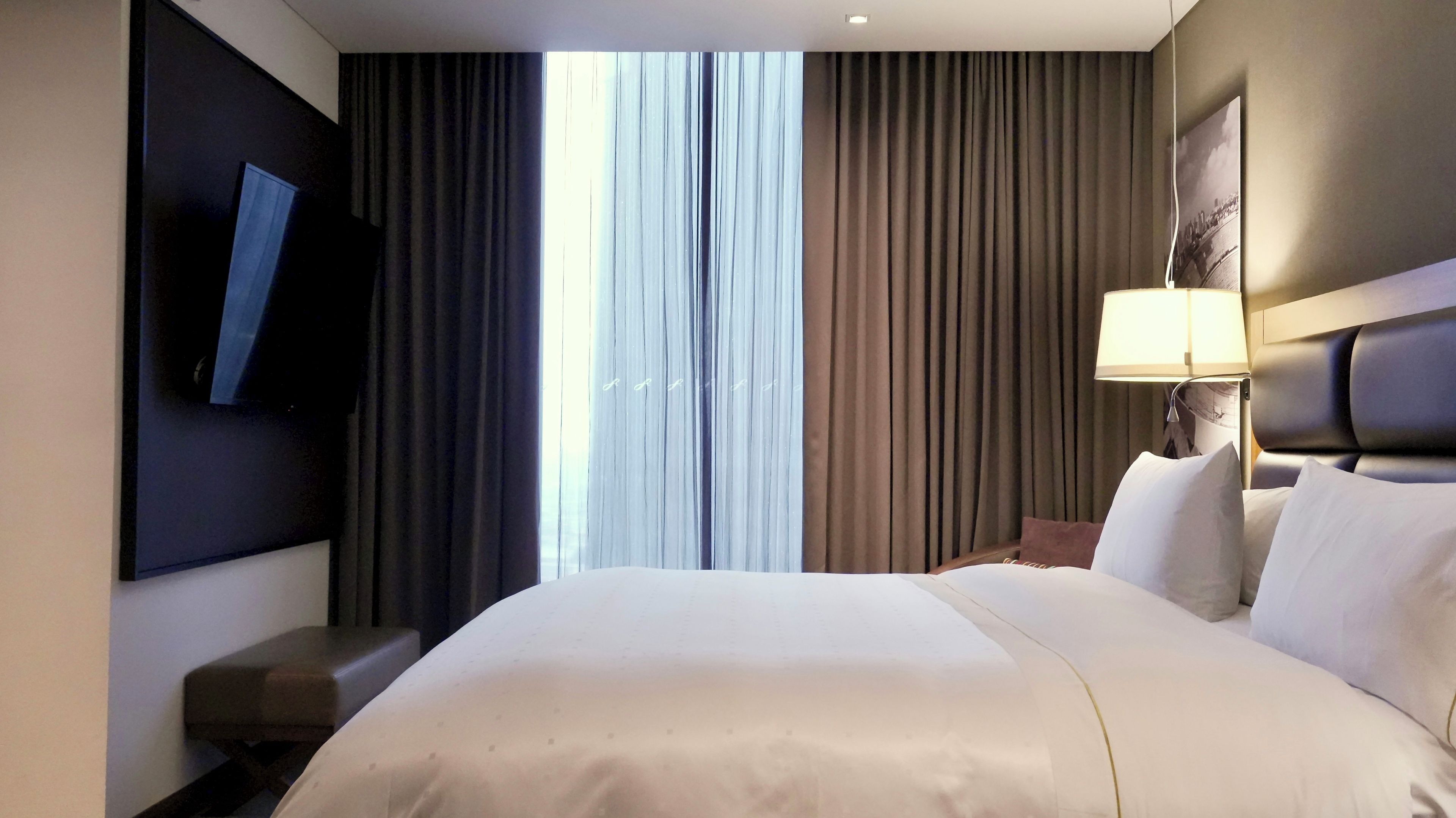 Une chambre d’hôtel avec un grand lit, un téléviseur à écran plat fixé au mur, une lampe de chevet et une fenêtre avec des rideaux.